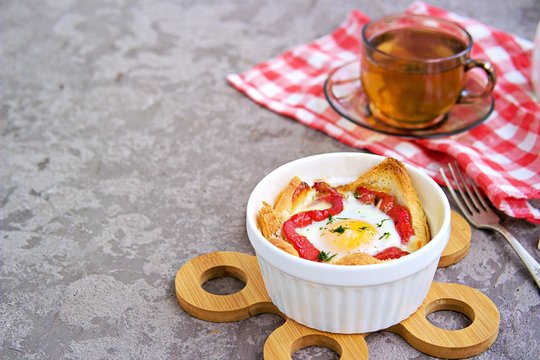 Breakfast, baked egg with cheese, sweet pepper and bread in a white portion form on a gray concrete background.