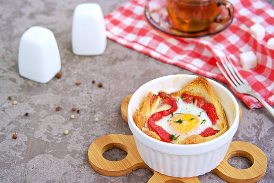 Breakfast, baked egg with cheese, sweet pepper and bread in a white portion form on a gray concrete background.