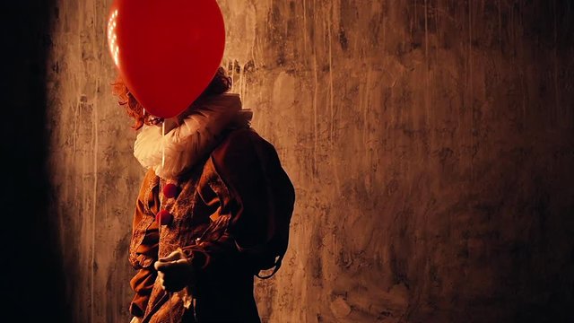 Creepy clown with colorful makeup in a carnival costume goes into the dark. Shooting in slow motion on dark red background