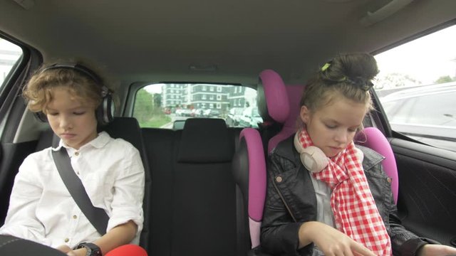 Brother and sister use the phone and tablet while traveling in the car. Boy and Girl with Headphones Playing a Tablet and smartphone in auto during ride