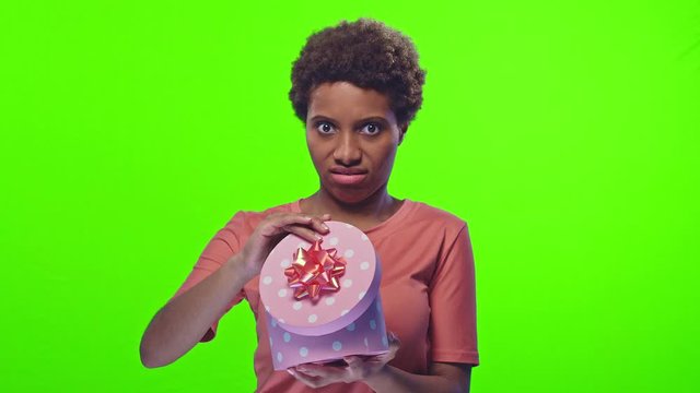 Positive emotional young pretty dark skinned woman happily opening a box with a gift and disappointed on a green background. Concept of surprise and unwarranted expectations. People and celebration