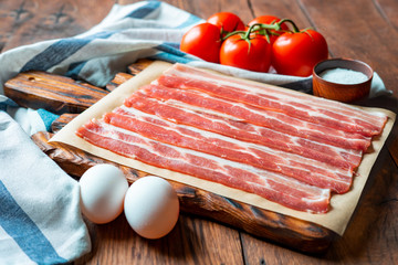 Raw bacon kitchen board, salt, tomatoes, eggs on a dark wooden background.