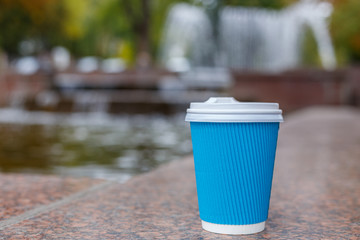 One blue paper coffee cup standing on granite surface in the city park on fountain background. Clouse up.