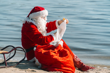 Obraz premium Santa Claus sits with a gift bag on the sandy shore of a lake and reads a list