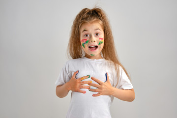 Little girl in white painted t-shirt is posing standing isolated on white and gesticulating with her colored in different paints palms and face. Art studio. Close-up.