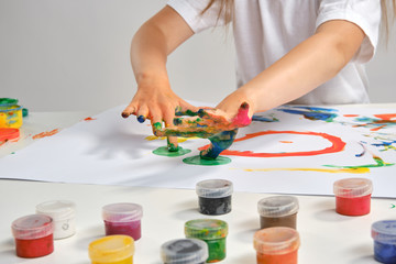 Little artist in white t-shirt standing at table with whatman and colorful paints, painting on it...