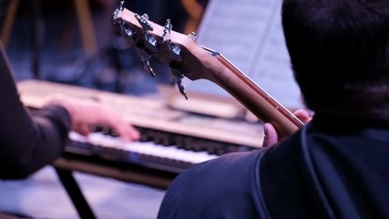A musician from the orchestra plays the bass guitar against the background of the hands of a pianist. A musical concert of classical or rock music. Close-up. Fingers fingering the strings. The view fr