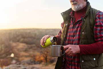 Eldery tourist man with gray hair and beard in plaid shirt drinking tea from a thermos to an iron...