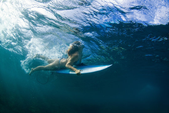 Adult active girl in bikini in action - surfer with surf board dive underwater under breaking big ocean wave.