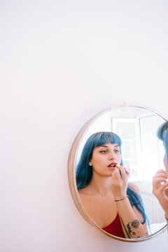 young woman with blue hair paints her lips in red in front of a mirror ?