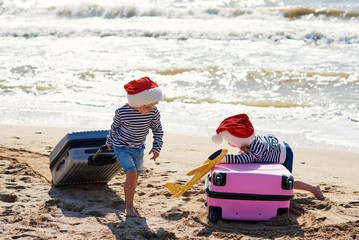 Children travelers. Little girls in Santa hats playing with toy airplane on sunny tropical beach. pulling a suitcase luggage. Christmas and new year family vacation, travel concept