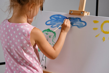 Little girl artist in a pink dress is standing behind easel and painting with brush on canvas at art studio with white walls. Medium close-up shot.