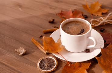 Cup of coffee with black coffee in a saucer on a wooden background, red fallen autumn leaves, cinnamon, a slice of dried orange