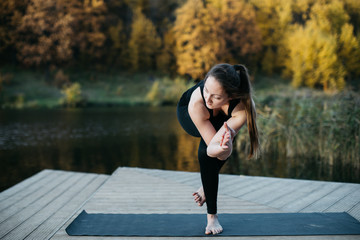 Young woman doing yoga asana in the nature with the lake view. Evening workout outdoors, sports and healthy lifestyle