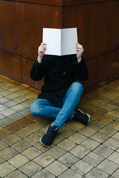 Man with book at face level sitting near building