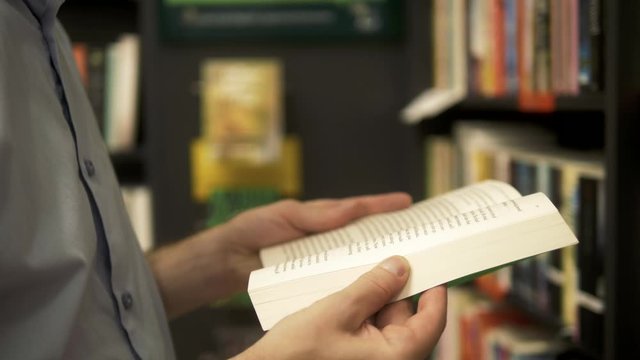 close-up of young caucasian man in a bookstore with an opened book in his hands reading something with other bookshelves in background