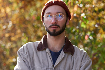 Portrait of attractive young student in hat and glasses with yellow autumn trees at background.