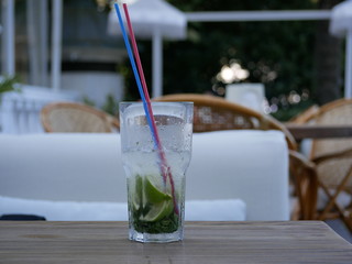 A glass of cold Mojito with ice is on the table on the summer veranda of the cafe. The glass with the tube is covered with droplets of condensate. Drink on a hot summer day.