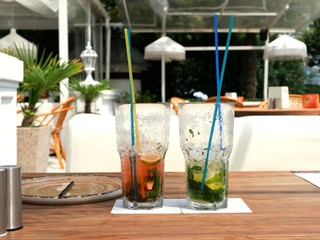 Two glasses of cold mojitos and an iced strawberry cocktail sit on a table on the cafe's summer veranda. Glasses with a tube covered with droplets of condensate. Refreshing drinks on a hot summer day.