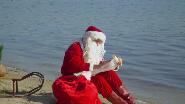 Santa Claus sits with a gift bag on the sandy shore of the lake and writes something down the list