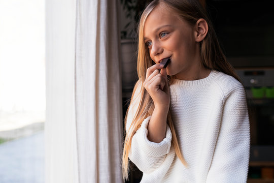 Positive girl eating chocolate near window with curtains