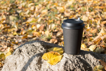A black paper Cup of hot coffee stands on a stone, against a blurred background of autumn yellow leaves