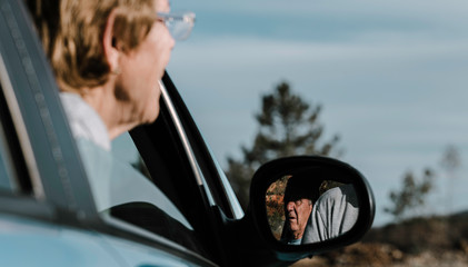 A old woman out of focus with her head out the window and focus on the elderly man reflected in the car mirror