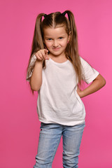 Little girl with ponytails, dressed in white t-shirt and blue jeans is posing against a pink studio background. Close-up shot. Sincere emotions.