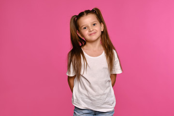 Little girl with ponytails, dressed in white t-shirt and blue jeans is posing against a pink studio background. Close-up shot. Sincere emotions.