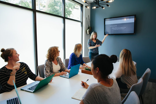 Woman In Conference Room Stands While Coworkers Sit At Table