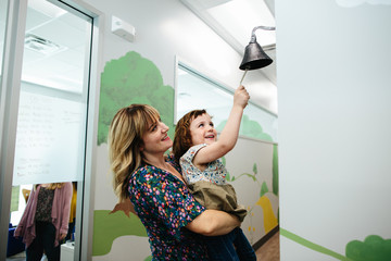 Teacher holds little girl up so that she can ring a bell in school