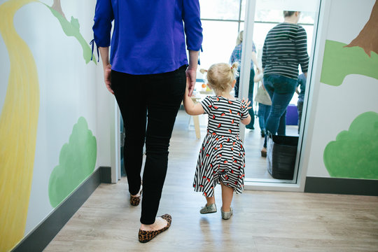 Toddler girl and mother hold hands while walking into a room at school