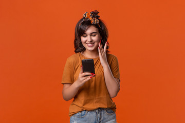 Pretty brunette woman in a t-shirt and beautiful headband checking smartphone for notifications isolated over orange background. Daily communication