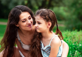 Beautiful playful mother embracing and talking with her cute angry daughter on summer green grass background in sunny day.