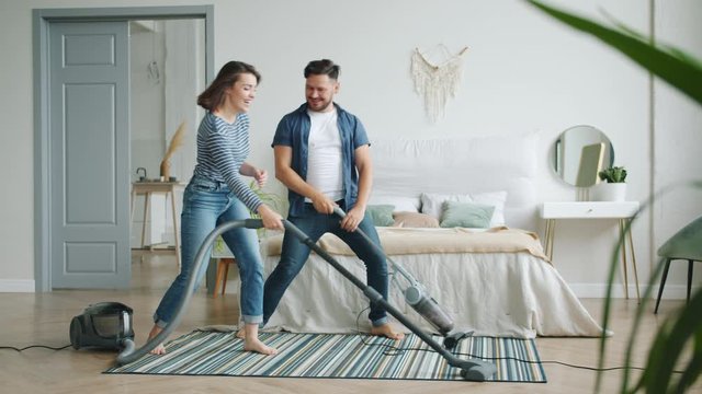 Girl and guy cheerful couple are vacuuming carpet in bedroom using 2 vacuum cleaners laughing having fun during clean-up at home. Family and housework concept.