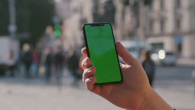 Man's hand showing vertical smartphone with mock-up chroma key greenscreen on outdoor background. Young person with modern iphone in busy city street close-up.