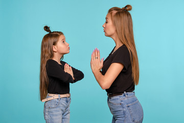 Mom and daughter with a funny hairstyles, dressed in black shirts and blue denim jeans are posing against a blue studio background. Close-up shot.