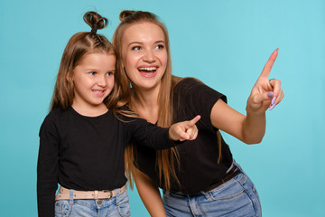 Mom and daughter with a funny hairstyles, dressed in black shirts and blue denim jeans are posing against a blue studio background. Close-up shot.