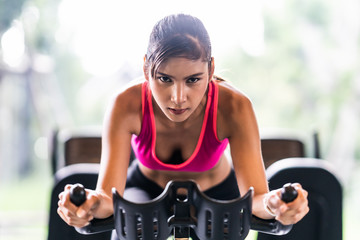Fototapeta na wymiar Beautiful Asian woman exercising on stationary cycling machine in indoor fitness gym, determination face. Sport recreational activity, people workout, or healthy lifestyle concept