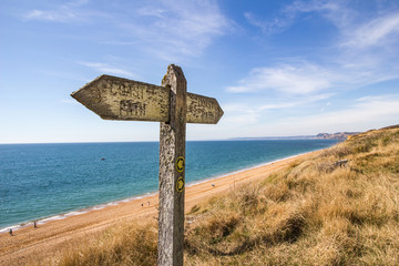 A weather worn wooden sign points the way of the west Dorset coast path along the section near Burton Bradstock and showing Chesil Beach and the shoreline - Powered by Adobe