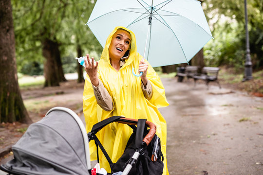 Beautiful And Happy Middle Age Woman With Her Baby Walking In City Park On Rainy Day.