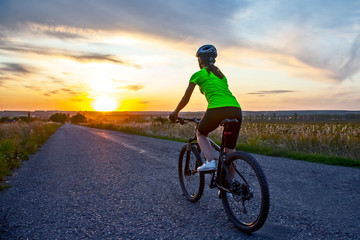 Naklejka na ściany i meble Beautiful girl cyclist rides a bicycle on the road in the sunset.
