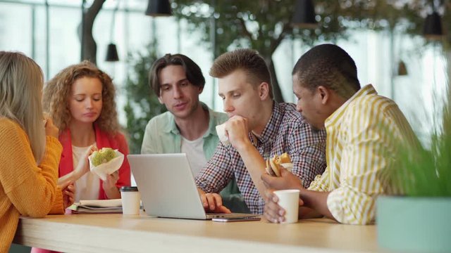 Group of diverse students biting burgers and browsing laptop while sitting at table and doing homework in cozy cafeteria