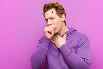 young red head man feeling ill with a sore throat and flu symptoms, coughing with mouth covered against purple wall
