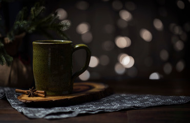 dark cozy space with mug of tea with cinnamon stick and star anise, on wooden surface with cloth napkin, small christmas tree background, holiday