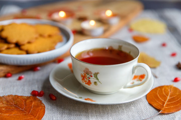 Cozy homemade tea party. Autumn composition. Porcelain cup of hot tea, Gingerbread cookie, burning candles on background, yellow fallen leaves, seeds pomegranate on a linen tablecloth. Soft focus. 