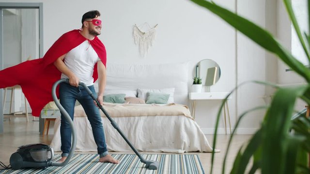 Slow motion of smiling man in super hero costume vacuuming floor in apartment using modern vacuum cleaner doing housework alone. People and household concept.