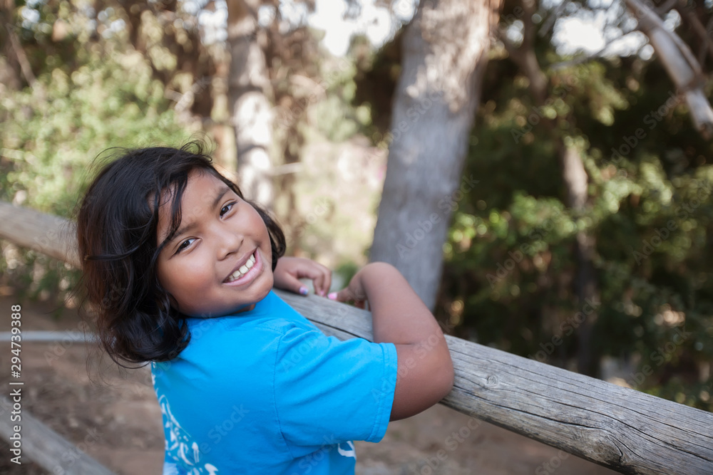 Wall mural a little girl with a big smile who is holding on to a wooden fence and surrounded by trees in a natu