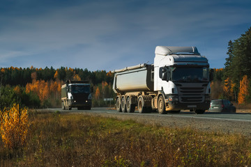 A large trucks are driving along a dirt road against a forest and blue sky.