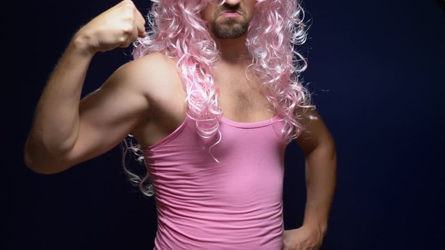 crazy handsome young guy in a curly wig and a pink t-shirt against a dark background is dancing funny, shows his muscles